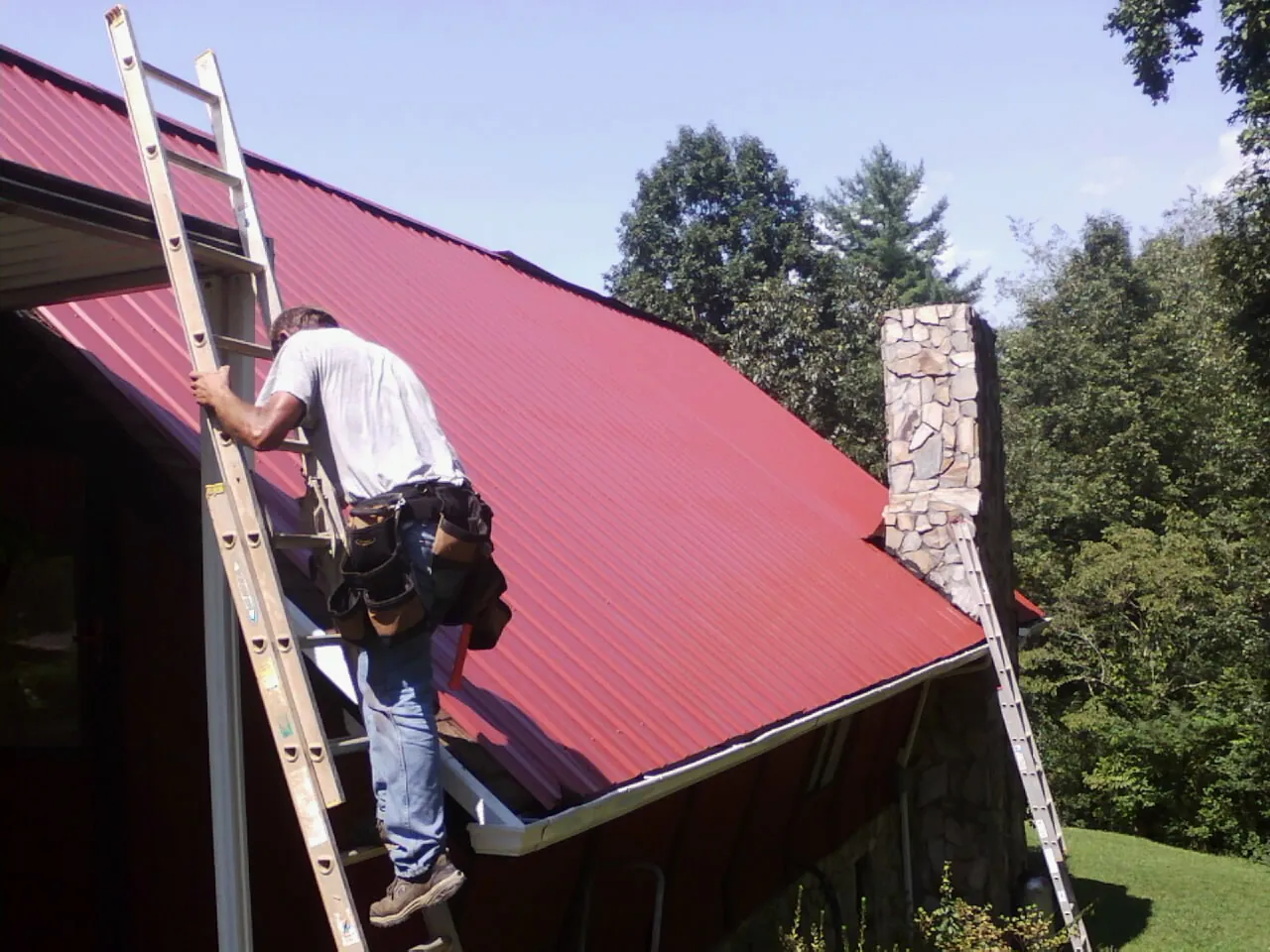 Roofer on a ladder beside a red metal roof and stone chimney — Soffit & Fascia Repair by Roofing Brothers in Minneapolis
